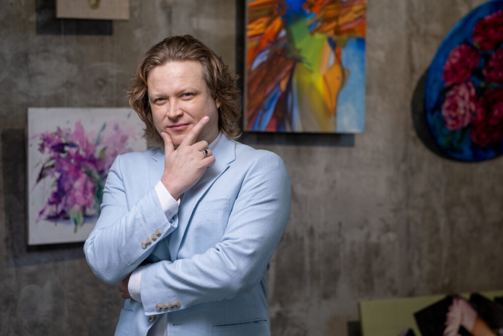Mature artist in blue jacket standing against wall with his artworks in art gallery and looking at you